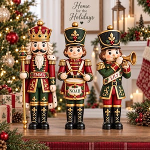 Puede incluir: Tres figuras decorativas de cascanueces con uniformes rojos y verdes. La figura de la izquierda es un rey que sostiene un bastón con el nombre EMMA. La figura central es un tamborilero con el nombre NOAH. La de la derecha es un trompetista con el nombre LILY.