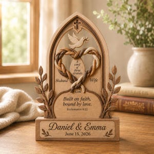 Könnte beinhalten: Eine hölzerne Bogenplatte mit einer geschnitzten Taube, einem geflochtenen Herz und dem Text A cord of three strands, Husband, Wife, God, Built on faith, bound by love, Ecclesiastes 4:12, Daniel & Emma und June 15, 2026, mit Blattverzierungen.
