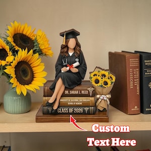 Könnte beinhalten: Eine Abschlussfigur einer Medizinstudentin in schwarzem Gewand und Mütze, die auf Büchern sitzt. Auf den Büchern steht "DR. SARAH J. REED", "CLASS OF 2026" und "UNIVERSITY OF CALIFORNIA, SAN FRANCISCO SCHOOL OF MEDICINE". Ein Strauß Sonnenblumen und eine Vase mit Sonnenblumen sind ebenfalls vorhanden.