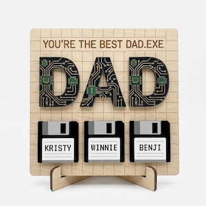 Może przedstawiać: Drewniana tablica z napisem "YOU'RE THE BEST DAD.EXE" i słowem "DAD" w projekcie obwodu drukowanego. Poniżej trzy dyskietki z imionami "KRISTY", "WINNIE" i "BENJI".