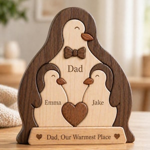 Peut inclure: Sculpture familiale en bois en forme de pingouins. Le mot "Dad" et les noms "Emma" et "Jake" sont gravés. Bordure marron foncé, centre en bois clair, un cœur et le texte "Dad, Our Warmest Place".