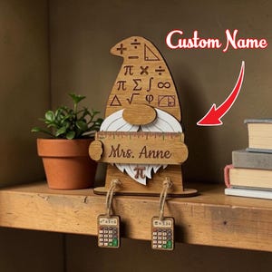 May include: Wooden gnome figurine with a hat featuring math symbols and a ruler beard. The name "Mrs. Anne" is engraved on the ruler. Two calculator charms hang from the gnome. A small plant in a terracotta pot is visible.