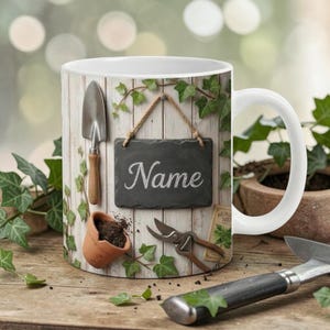 Può includere: Tazza in ceramica bianca con un design a tema giardino. Il design include una piccola lavagna con la parola "Name", attrezzi da giardinaggio, un piccolo vaso con terra e edera. La tazza è decorata con uno sfondo di assi di legno.