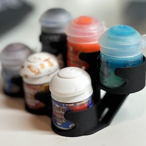 Puede incluir: Un soporte negro impreso en 3D para seis botes de pintura pequeños de varios colores. Los botes de pintura tienen tapas blancas, grises, naranjas y azules. El soporte está diseñado para mantener los botes de pintura organizados.