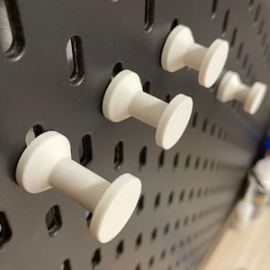 May include: Close-up of a black pegboard with several white pegboard hooks inserted. The hooks have a cylindrical body and a flat, circular head. The pegboard is designed for organizing tools and accessories.