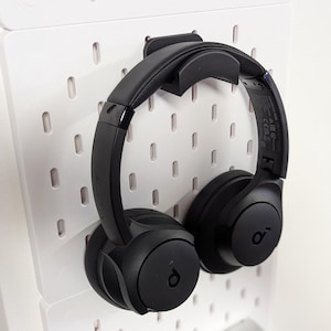 May include: Black over-ear headphones suspended on a white pegboard. The headphones feature a padded headband and earcups. The pegboard has a grid of holes for storage and organisation.