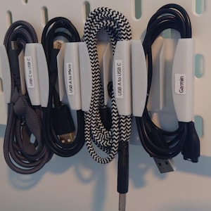 Op de afbeelding: Diverse USB-kabels, waaronder USB-C naar USB-C, USB A naar Micro en USB A naar USB-C, netjes georganiseerd met witte kabelhouders. Eén kabel is gemarkeerd met "Garmin". De kabels zijn in verschillende kleuren, waaronder zwart, grijs en een zwart-wit zigzagpatroon.