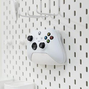 May include: A white Xbox controller with black thumbsticks and coloured buttons is mounted on a white pegboard. The pegboard has various white hooks and storage containers attached, creating a tidy gaming setup.