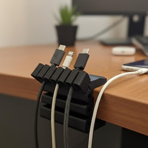 Puede incluir: Un organizador de cables negro con múltiples ranuras, que sujeta varios cables de carga. Un smartphone se está cargando en un escritorio de madera. El organizador está diseñado para mantener los cables ordenados.