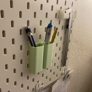May include: A light green pen holder with two compartments, holding pens of various colours. The holder is attached to a white pegboard, with a metal ruler and other accessories. The pegboard has evenly spaced oval holes.