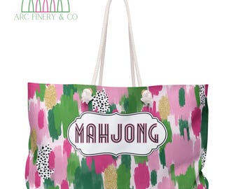 May include: A large tote bag with a colorful abstract design in pink, green, and gold. The bag features a white rope handle and the word "MAHJONG" in a decorative frame. The brand name "ARC FINERY & CO" is printed in green.