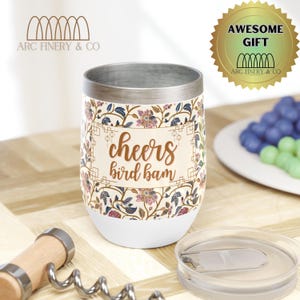 May include: White stainless steel wine tumbler with a floral pattern and the text "cheers bird bam" in gold lettering. The tumbler has a lid and is sitting on a wooden surface.