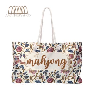May include: A white canvas tote bag with a floral pattern and the word "mahjong" printed in brown on the front. The bag has white rope handles.