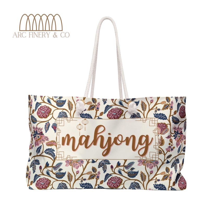 May include: A white canvas tote bag with a floral pattern and the word "mahjong" printed in brown on the front. The bag has white rope handles.