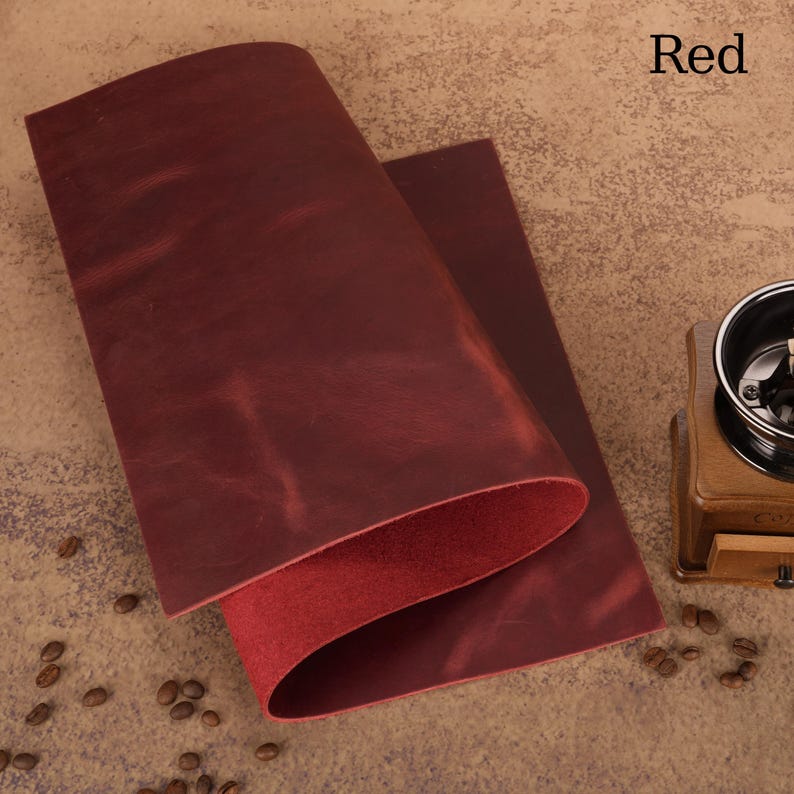 May include: A folded piece of red leather with a smooth, textured surface. The word "Red" is printed in white in the upper right corner. The leather is a deep, rich shade of red, with a slightly darker interior. Coffee beans and a coffee grinder are in the background.