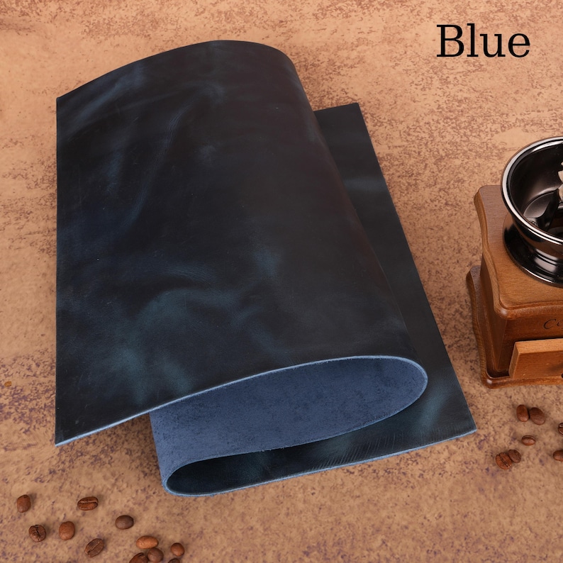 May include: Two sheets of blue leather with a distressed finish. The leather has a dark blue hue with lighter blue and green undertones. The word "Blue" is printed in white in the upper right corner. A wooden coffee grinder is in the background.