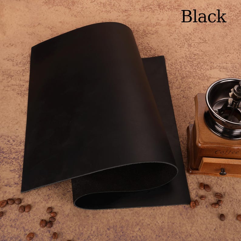 May include: Two sheets of black leather are displayed, with the word "Black" in the upper right corner. The leather has a smooth, matte finish. A wooden coffee grinder and coffee beans are in the background.
