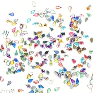 May include: A large assortment of colorful teardrop-shaped rhinestones, scattered on a white background. The rhinestones are iridescent and have a variety of colors, including pink, blue, green, yellow, and purple.