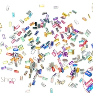 May include: Assortment of colorful, rectangular, iridescent rhinestones scattered on a white background.  The rhinestones are various shades of pink, blue, green, yellow, and orange.  The rhinestones are small and have a flat back.  The rhinestones are perfect for adding sparkle to crafts, nail art, and other projects.