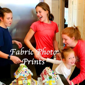 May include: A group of four people are gathered around two gingerbread houses decorated with colorful candies and frosting. The text "Fabric Photo Prints" is overlaid on the image. The people are smiling and laughing.