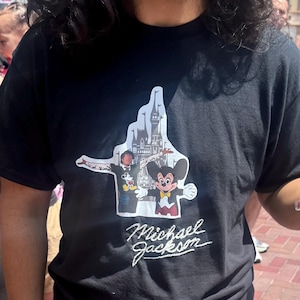 Può includere: Una t-shirt nera a maniche corte con una stampa grafica di Michael Jackson e Topolino davanti alla sagoma di un castello. Sotto la grafica, il nome Michael Jackson è stampato in un carattere corsivo bianco.