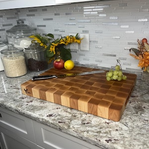 May include: A large wooden chopping board with a chequered pattern. The board is made of light and dark wood and has a natural finish. It is sitting on a granite worktop with a knife and some fruit and grapes on top.