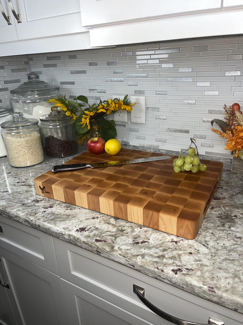 May include: A large wooden chopping board with a chequered pattern. The board is made of light and dark wood and has a natural finish. It is sitting on a granite worktop with a knife and some fruit and grapes on top.