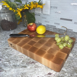 May include: A large, rectangular, wooden chopping board with a checkered pattern. The board is made of light wood and has a natural finish. There are some grapes, a lemon, and an apple on the board. A silver knife is also on the board.