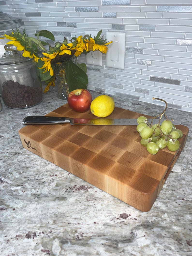 May include: A large, rectangular, wooden chopping board with a checkered pattern. The board is made of light wood and has a natural finish. There are some grapes, a lemon, and an apple on the board. A silver knife is also on the board.
