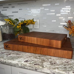 May include: Two wooden cutting boards stacked on top of each other on a granite countertop. The boards are both brown and have a bird logo engraved on them.