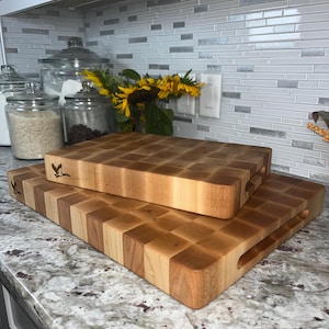 May include: Two wooden chopping boards, one stacked on top of the other. The top board is smaller and has a handle. Both boards are made of light wood with a chequered pattern.