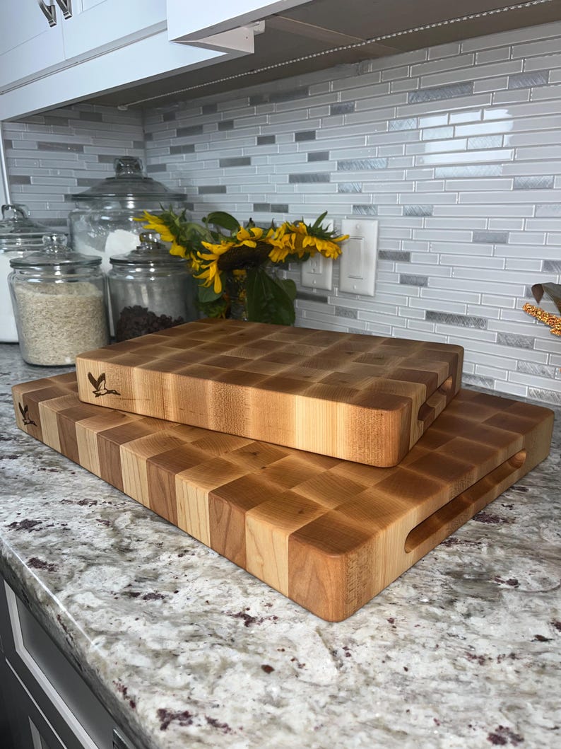 May include: Two wooden chopping boards, one stacked on top of the other. The top board is smaller and has a handle. Both boards are made of light wood with a chequered pattern.
