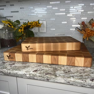 May include: Two wooden cutting boards stacked on top of each other on a granite countertop. The top board is a lighter wood with a checkered pattern and the bottom board is a darker wood with a checkered pattern. Both boards have a bird design engraved on them.