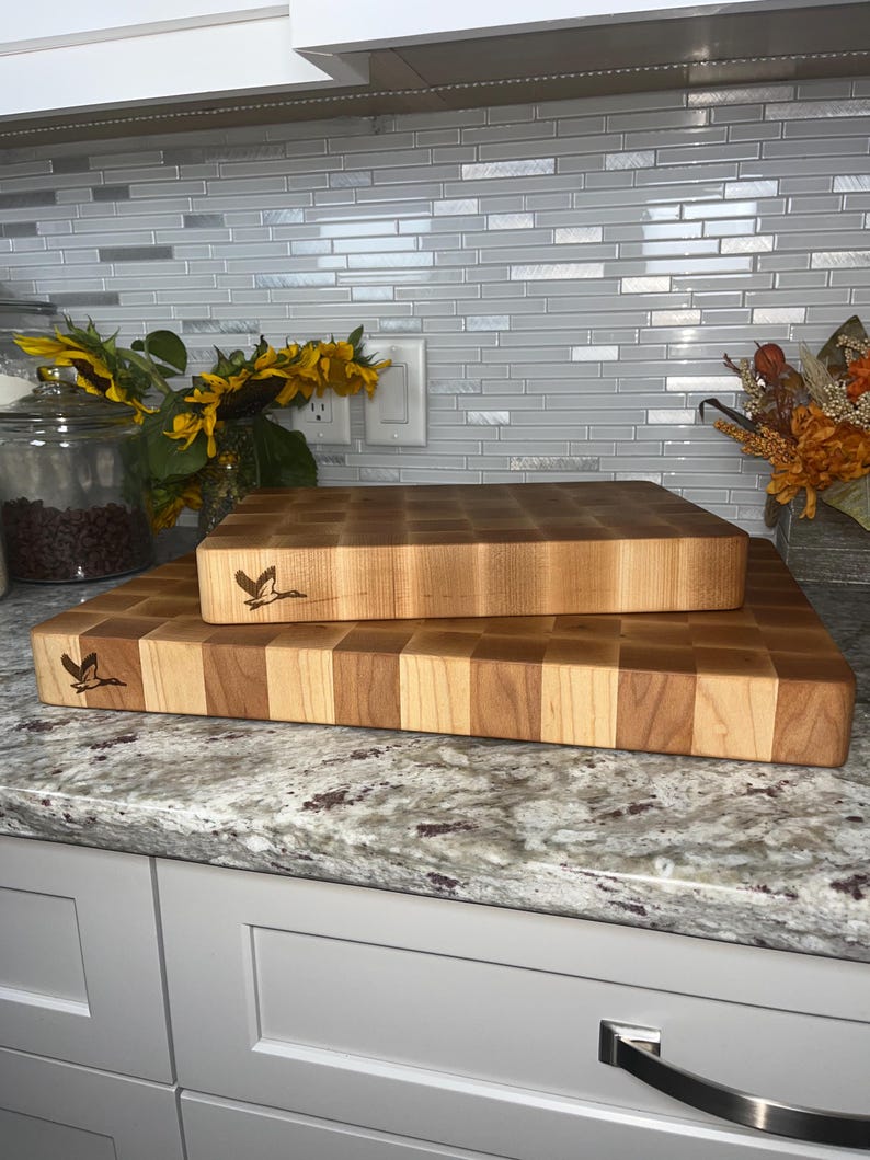 May include: Two wooden chopping boards stacked on top of each other on a granite worktop. The top board is a lighter wood with a chequered pattern and the bottom board is a darker wood with a chequered pattern. Both boards have a bird design engraved on them.