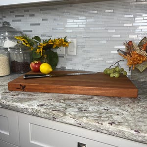 May include: A large, rectangular, wooden cutting board with a light brown finish. The board has a small, engraved bird design in the upper left corner. A silver knife is resting on the board. The board is sitting on a white countertop with a granite pattern.