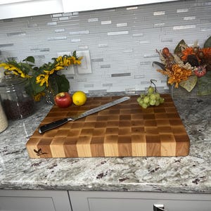 May include: A large, rectangular, wooden chopping board with a chequered pattern. The board is made of light and dark wood, and has a small, engraved design of a bird on one end. The board is sitting on a granite worktop.