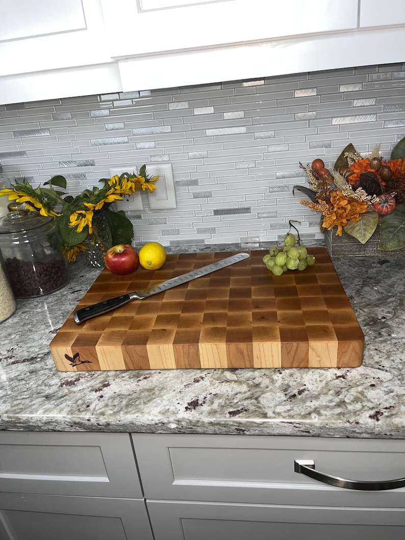 May include: A large, rectangular, wooden chopping board with a chequered pattern. The board is made of light and dark wood, and has a small, engraved design of a bird on one end. The board is sitting on a granite worktop.