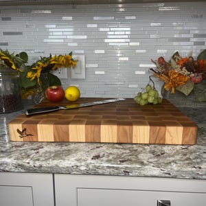 May include: A large, rectangular, wooden chopping board with a chequered pattern in shades of brown and light wood. The board has a small, engraved bird design in the top left corner. A knife and some fruit are on the board.