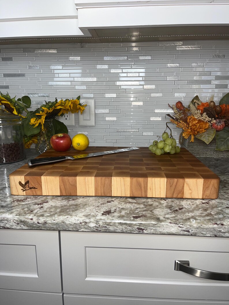 May include: A large, rectangular, wooden chopping board with a chequered pattern in shades of brown and light wood. The board has a small, engraved bird design in the top left corner. A knife and some fruit are on the board.