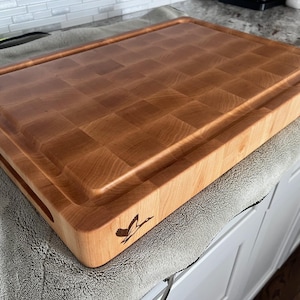May include: A large, rectangular, wooden chopping board with a raised edge and a juice groove. The board is made of maple wood and has a checkered pattern. The board has a small, engraved logo on the side.