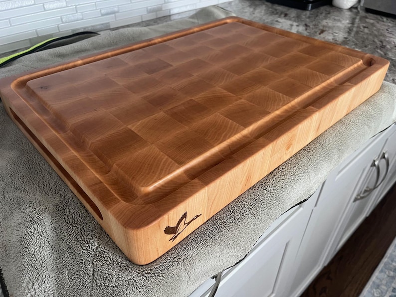 May include: A large, rectangular, wooden chopping board with a raised edge and a juice groove. The board is made of maple wood and has a checkered pattern. The board has a small, engraved logo on the side.