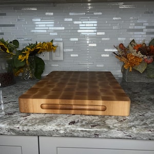 May include: A large, rectangular, wooden chopping board with a handle. The board is made of light wood and has a checkered pattern. The board is sitting on a granite worktop.