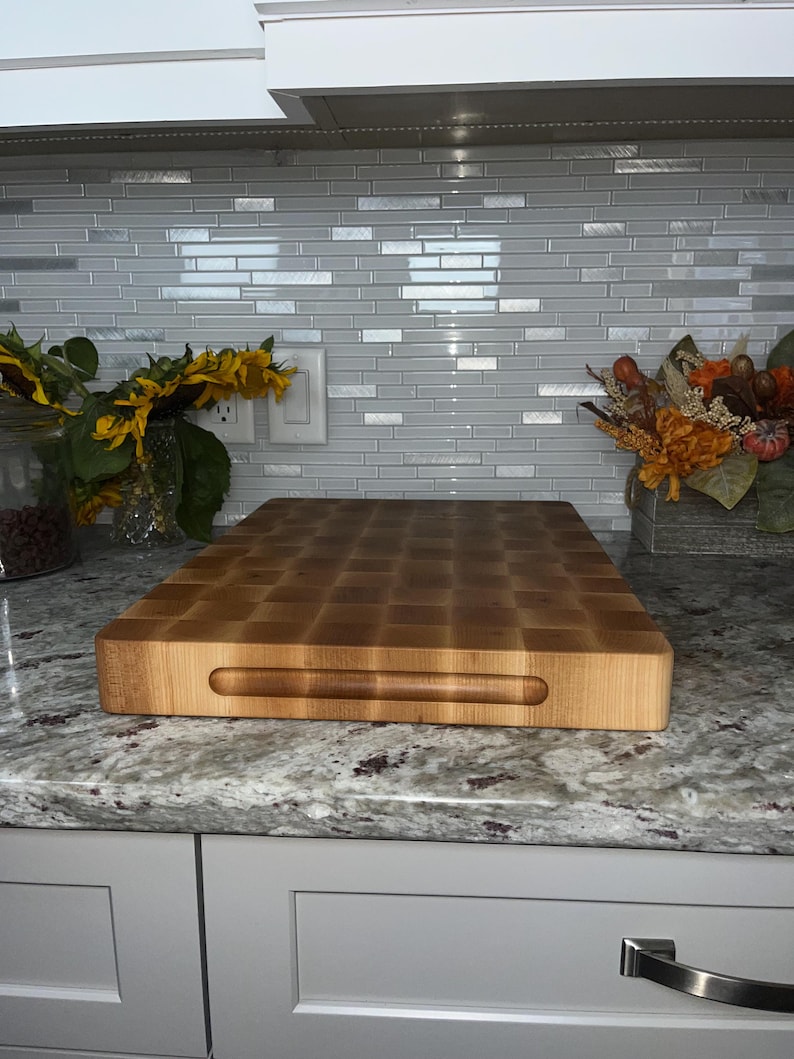May include: A large, rectangular, wooden chopping board with a handle. The board is made of light wood and has a checkered pattern. The board is sitting on a granite worktop.