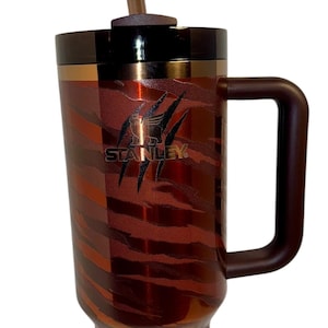以下が含まれることがあります： 茶色のハンドルと装飾的なストローが付いたスタンレーのトラベルマグ。マグカップは、茶色とゴールドの色合いのトラ柄模様です。スタンレーのロゴが見えます。ストローには装飾的なリボンが付いています。