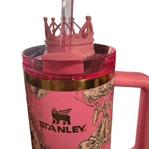 Könnte beinhalten: Rosa Stanley-Becher mit kronenförmigem Deckel und klarem Strohhalm. Der Becher hat ein goldenes Band und einen rosa Griff. Der Becher hat Illustrationen von Schmetterlingen und Figuren auf rosa Hintergrund. Das Stanley-Logo ist sichtbar.