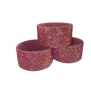 May include: Three pink, cylindrical napkin rings. Each ring is covered in small, sparkling rhinestones. The napkin rings are arranged in a triangular pattern, with one ring on top of the other two.