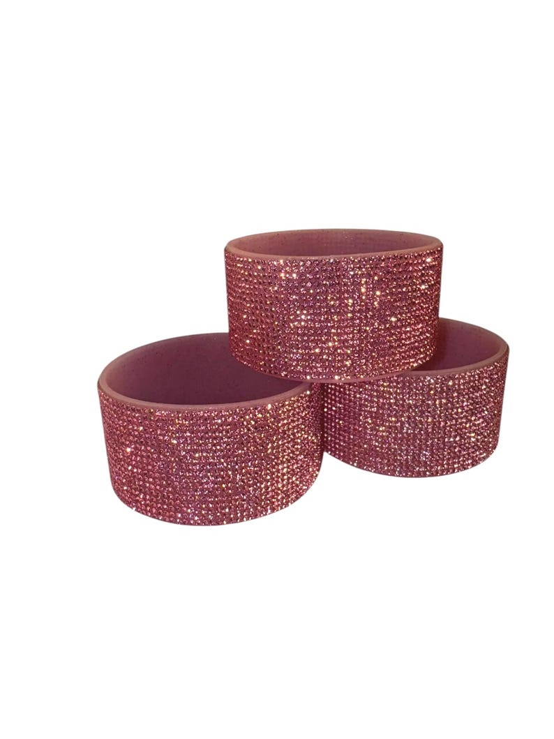 May include: Three pink, cylindrical napkin rings. Each ring is covered in small, sparkling rhinestones. The napkin rings are arranged in a triangular pattern, with one ring on top of the other two.
