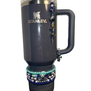 Op de afbeelding: Een grijze Stanley-reismok met een zwarte handgreep en een zilveren logo. De mok heeft een kralenbedel aan de handgreep en een stapel kleurrijke kralenarmbanden rond de onderkant.