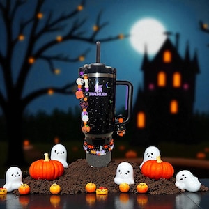 May include: A black Stanley tumbler with a handle, decorated with Halloween-themed charms and a beaded bracelet. The tumbler features the Stanley logo and a starry night design. The scene includes pumpkins, ghosts, and a spooky background.