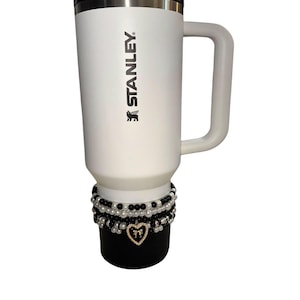 Op de afbeelding: Witte Stanley reismok met een zwarte deksel en handvat. De mok heeft het Stanley-logo in zwart gedrukt. De basis van de mok is versierd met meerdere strengen zwarte en witte kralen en een gouden hartbedel.